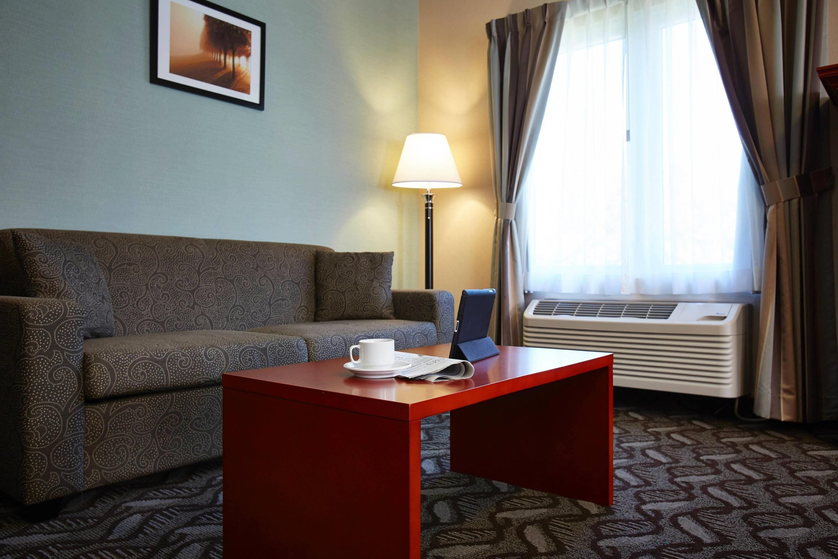 Ett hotellrum med en soffa, ett litet bord med en kopp och en bok, en lampa och en luftkonditioneringsanläggning.