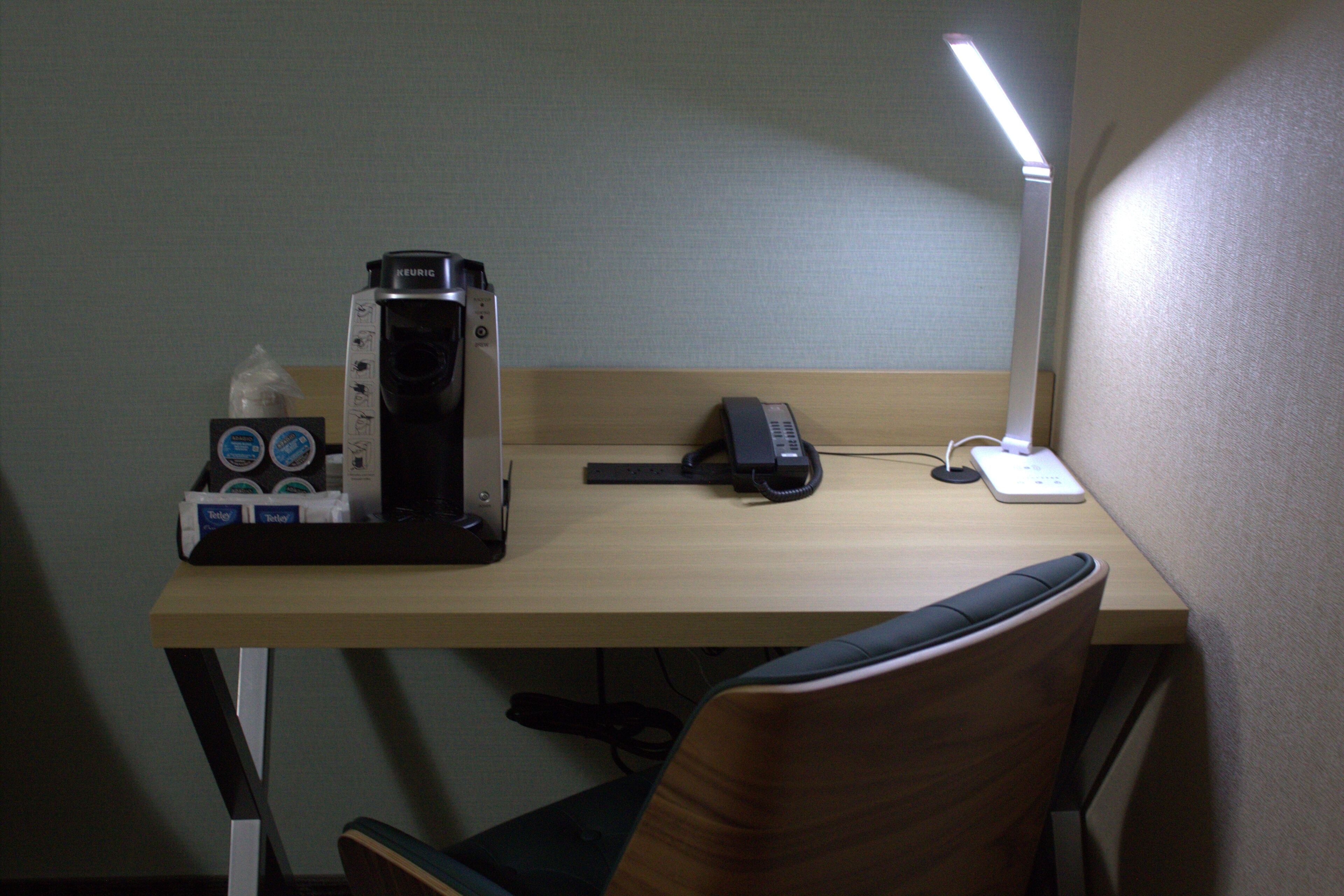 A hotel room with a desk, a coffee maker, a telephone, and a desk lamp.