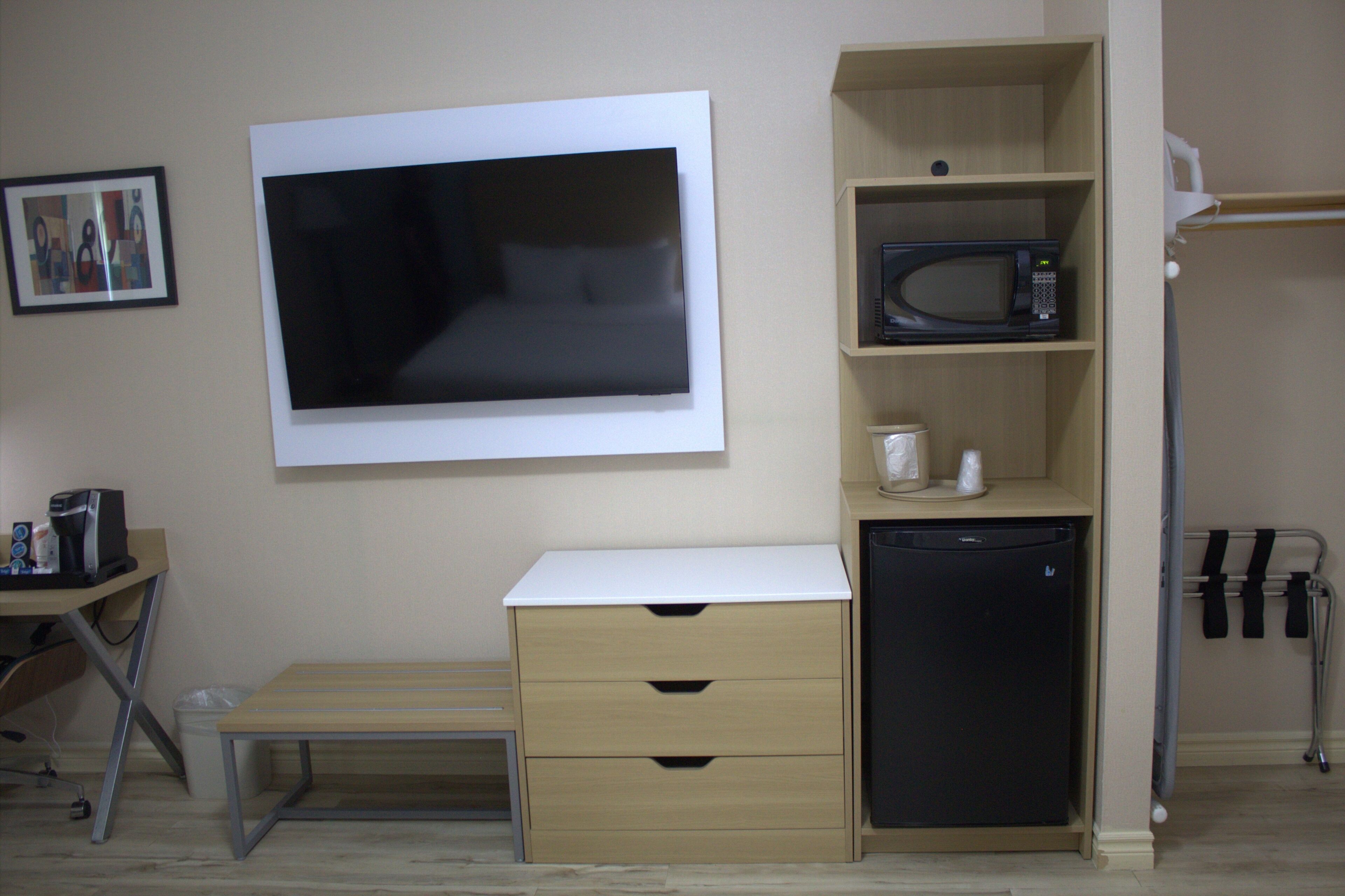 A compact room with a bed, bedside table, a mounted TV, a microwave, a small refrigerator, and a shelf with a microwave on it.