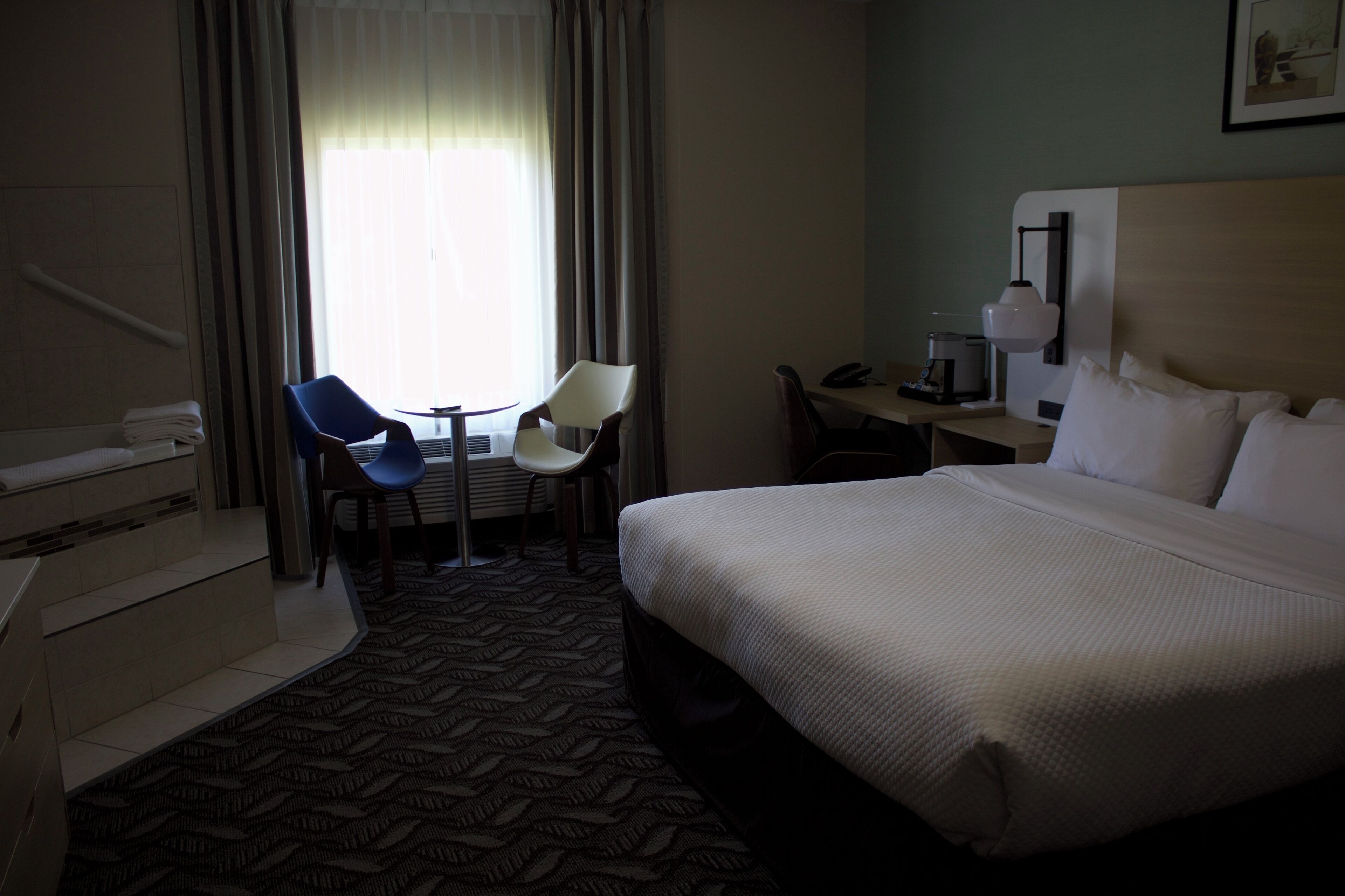 A hotel room with a bed, a desk, two chairs, a coffee maker, and a window with curtains.
