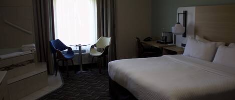 A hotel room with a bed, a desk, two chairs, a coffee maker, and a window with curtains.
