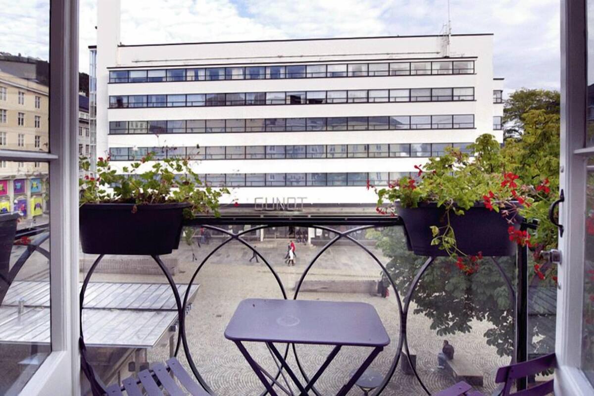 A balcony with two tables and two hanging planters with flowers, overlooking a building with the sign "GUNOT".