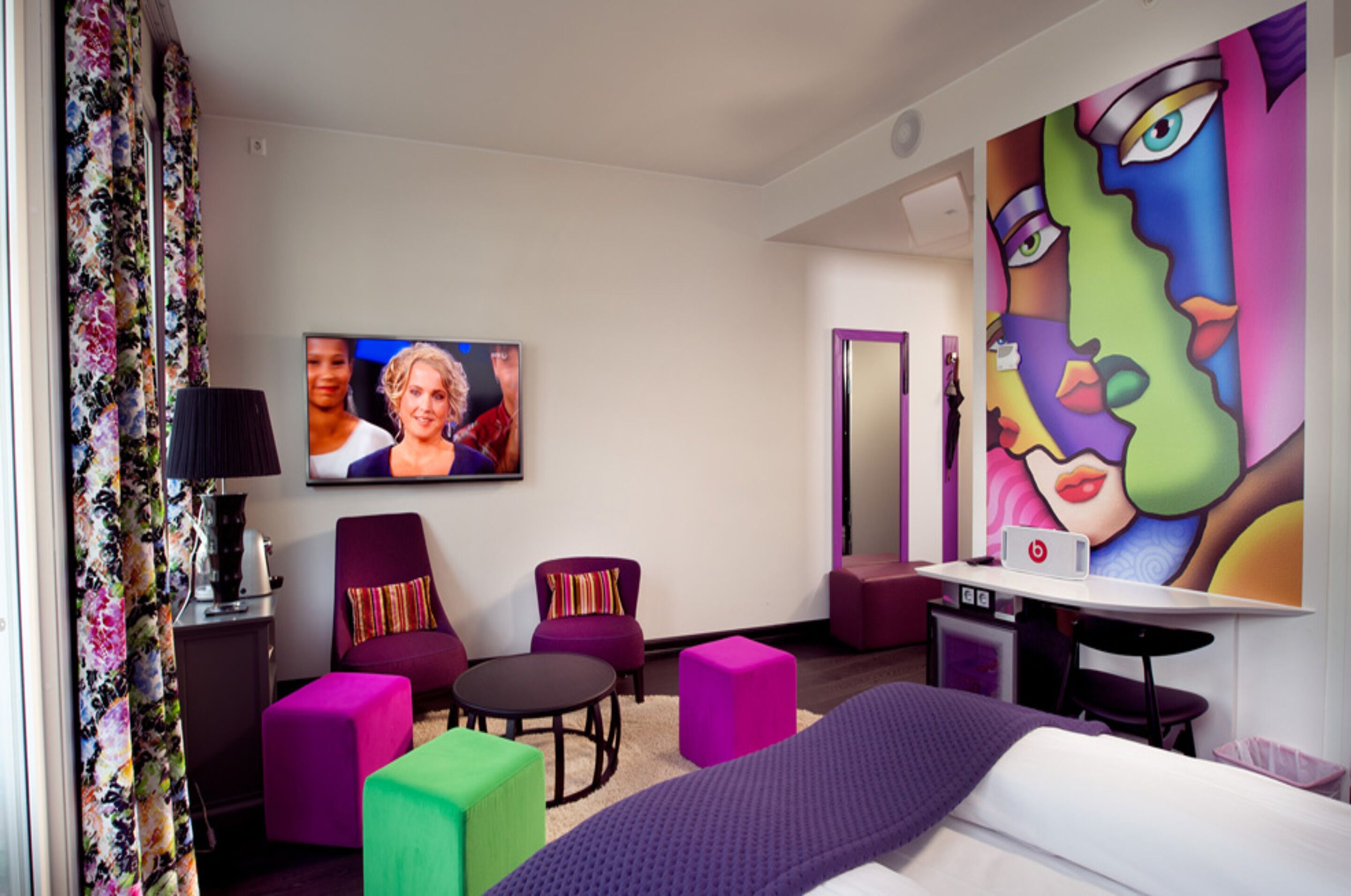 A hotel room with a bed, a desk, a chair, a small table, and colorful artwork on the walls.