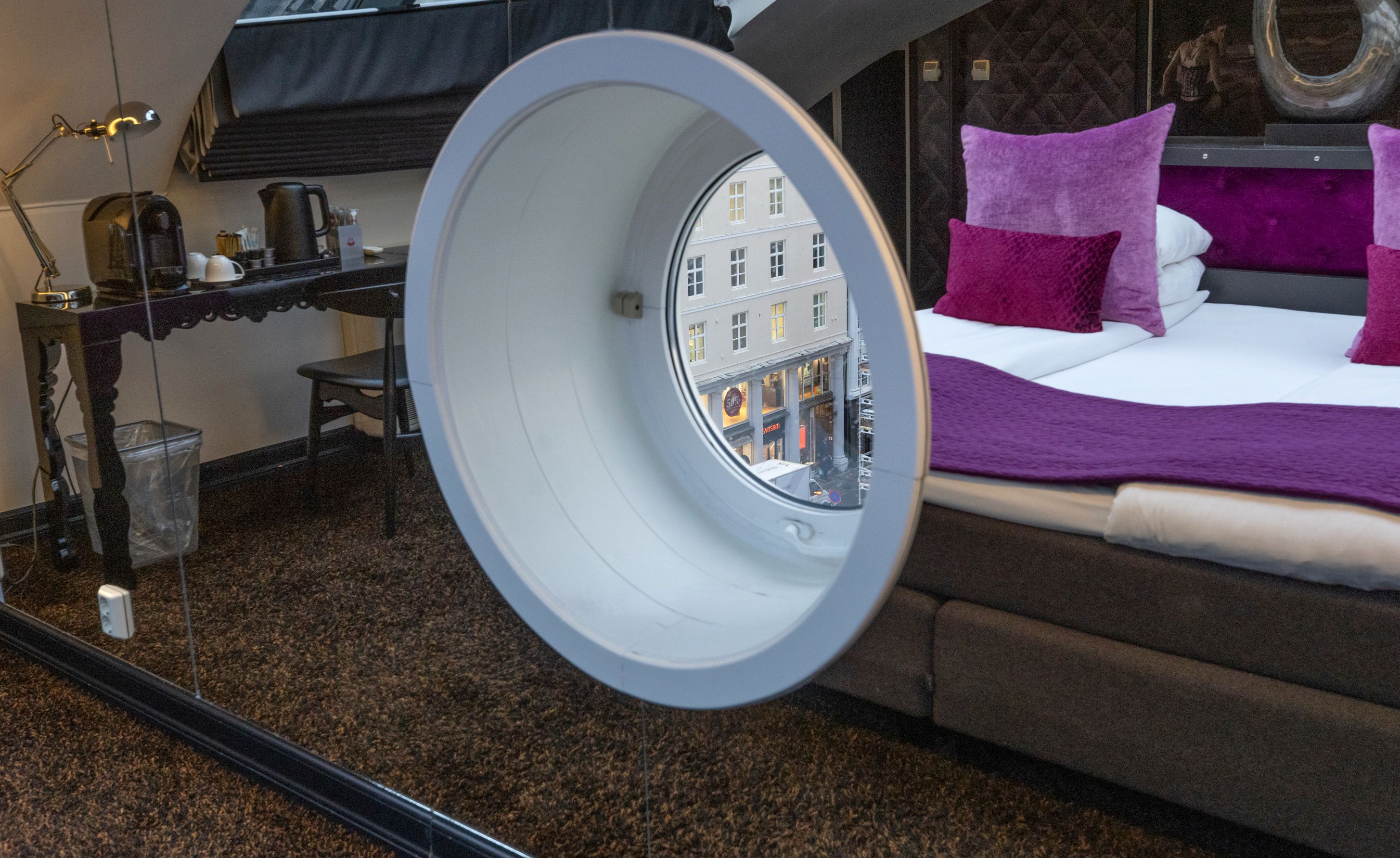 Une chambre d’hôtel avec un lit, un bureau et une vue sur les bâtiments, visible à travers une fenêtre circulaire.