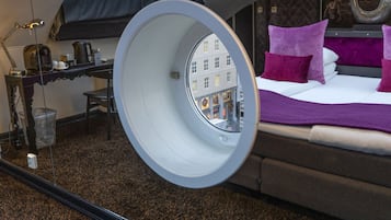 Une chambre d’hôtel avec un lit, un bureau et une vue sur les bâtiments, visible à travers une fenêtre circulaire.
