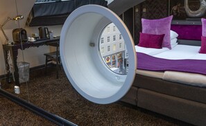 A hotel room with a bed, a desk, and a view of buildings through a circular window.