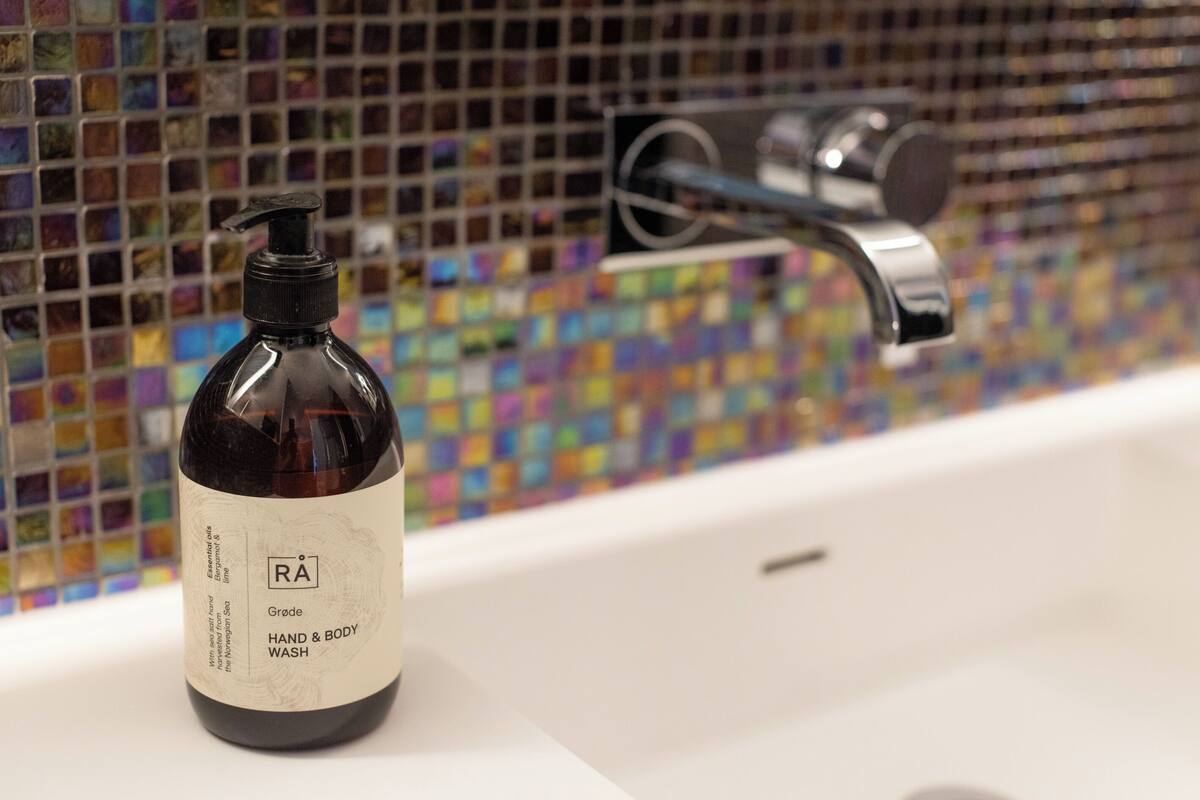 A bottle of hand and body wash on a bathroom sink with a colorful mosaic tile backsplash.