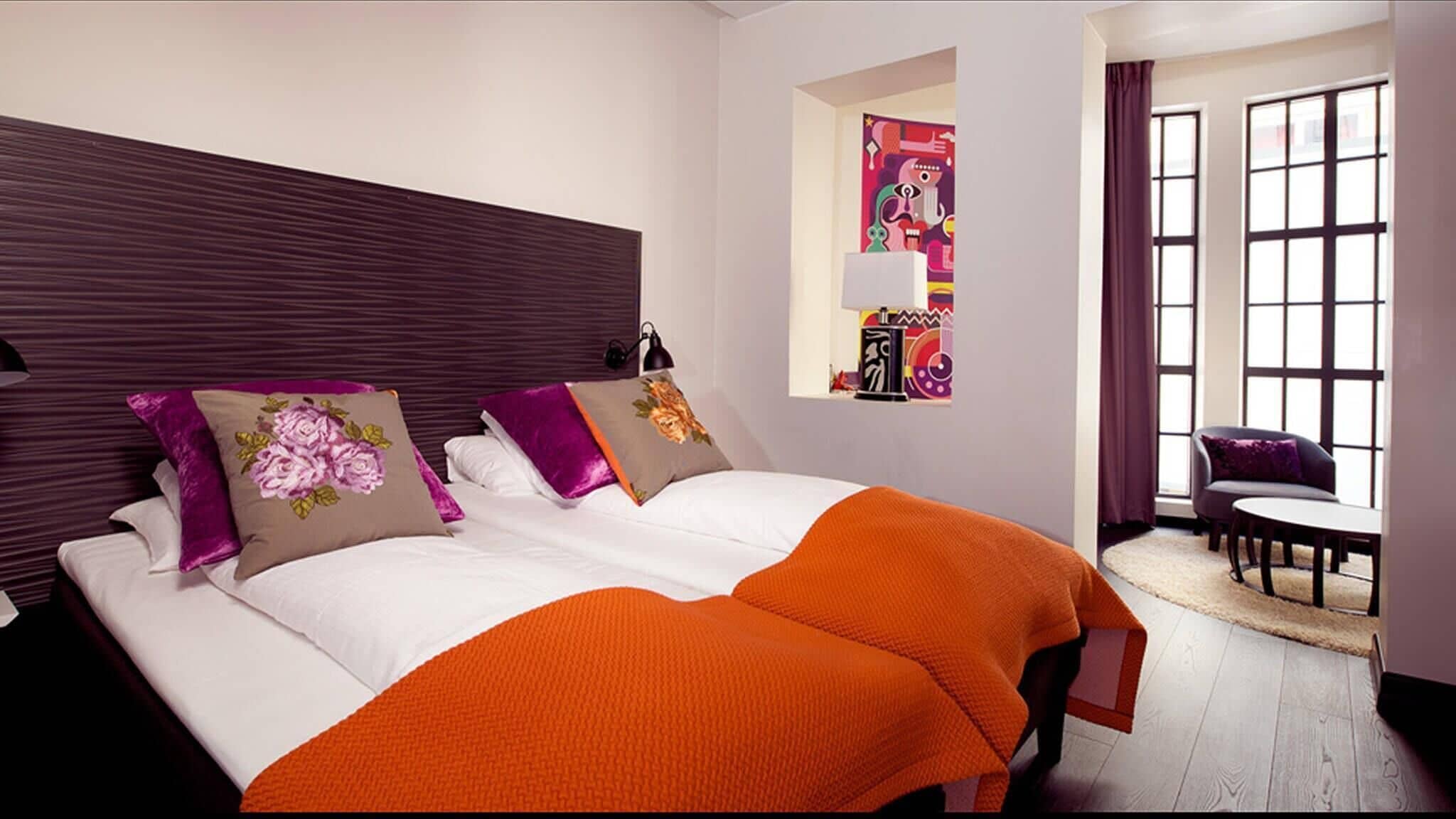 A hotel room with a large bed, two pillows, a nightstand, a lamp, a chair, and a window with curtains.