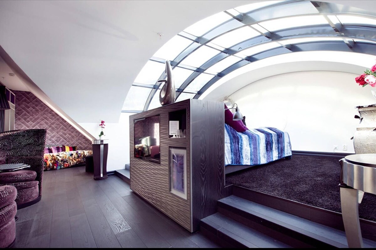 A modern interior with a curved ceiling, a bed with a striped blanket, a small table, and a seating area with cushions.