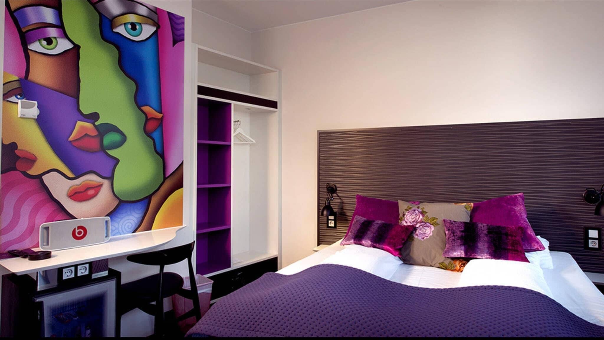 A modern bedroom with a large bed, purple bedding, and a vibrant abstract painting on the wall.