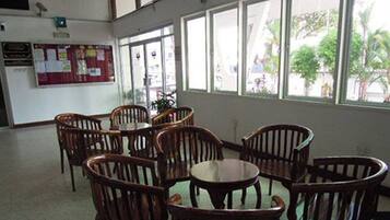 A room with wooden chairs and a table, large windows with a view of greenery outside, and a bulletin board on the wall.