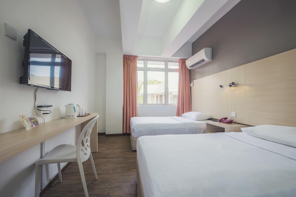 A hotel room with two beds, a desk with a flat-screen TV, a chair, a window with curtains, and an air conditioning unit.