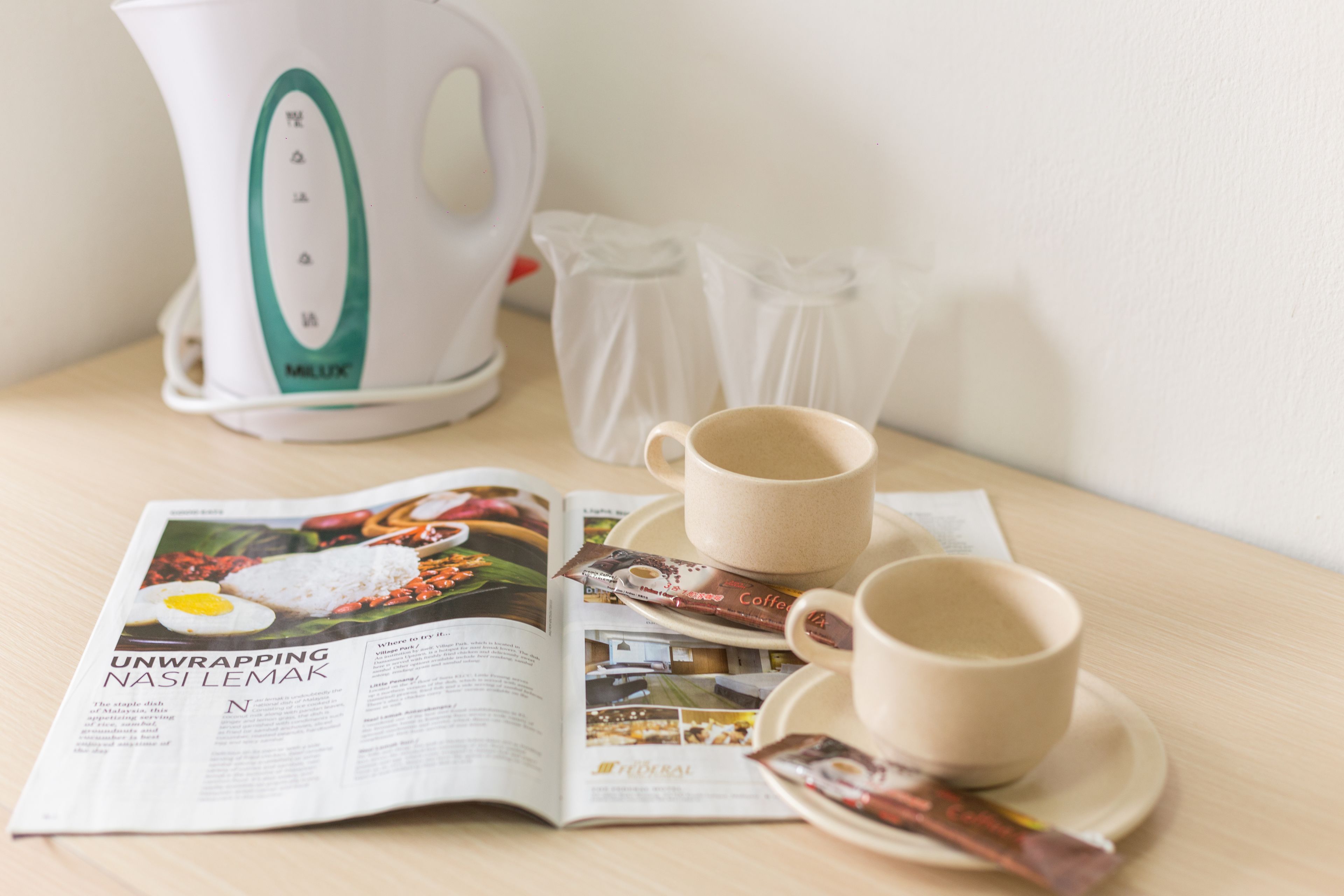 A magazine on a table with a coffee cup, saucer, and a white kettle.