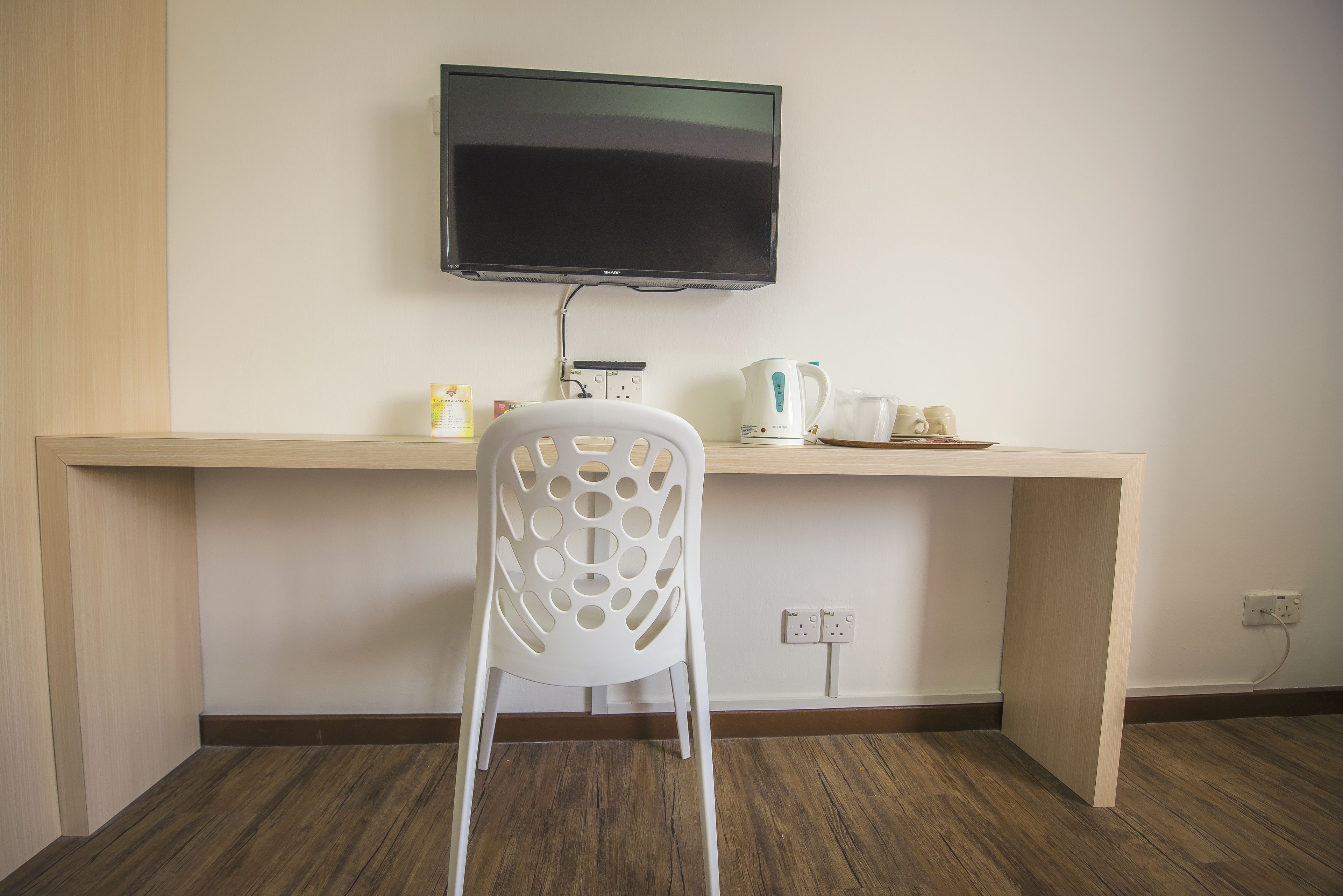 A room with a wooden desk, a white plastic chair, a flat-screen TV mounted on the wall, and a coffee maker on the desk.