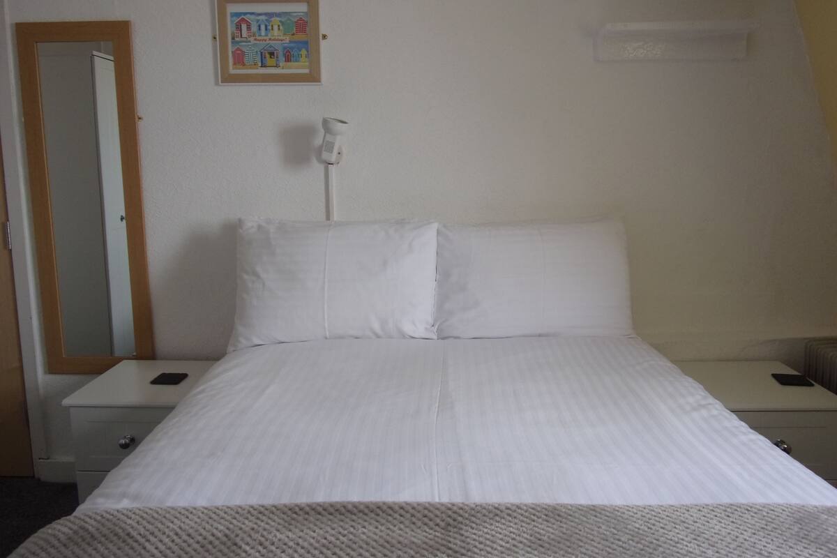 A neatly made bed with white linens in a simple room with a wooden door and a small bedside table.