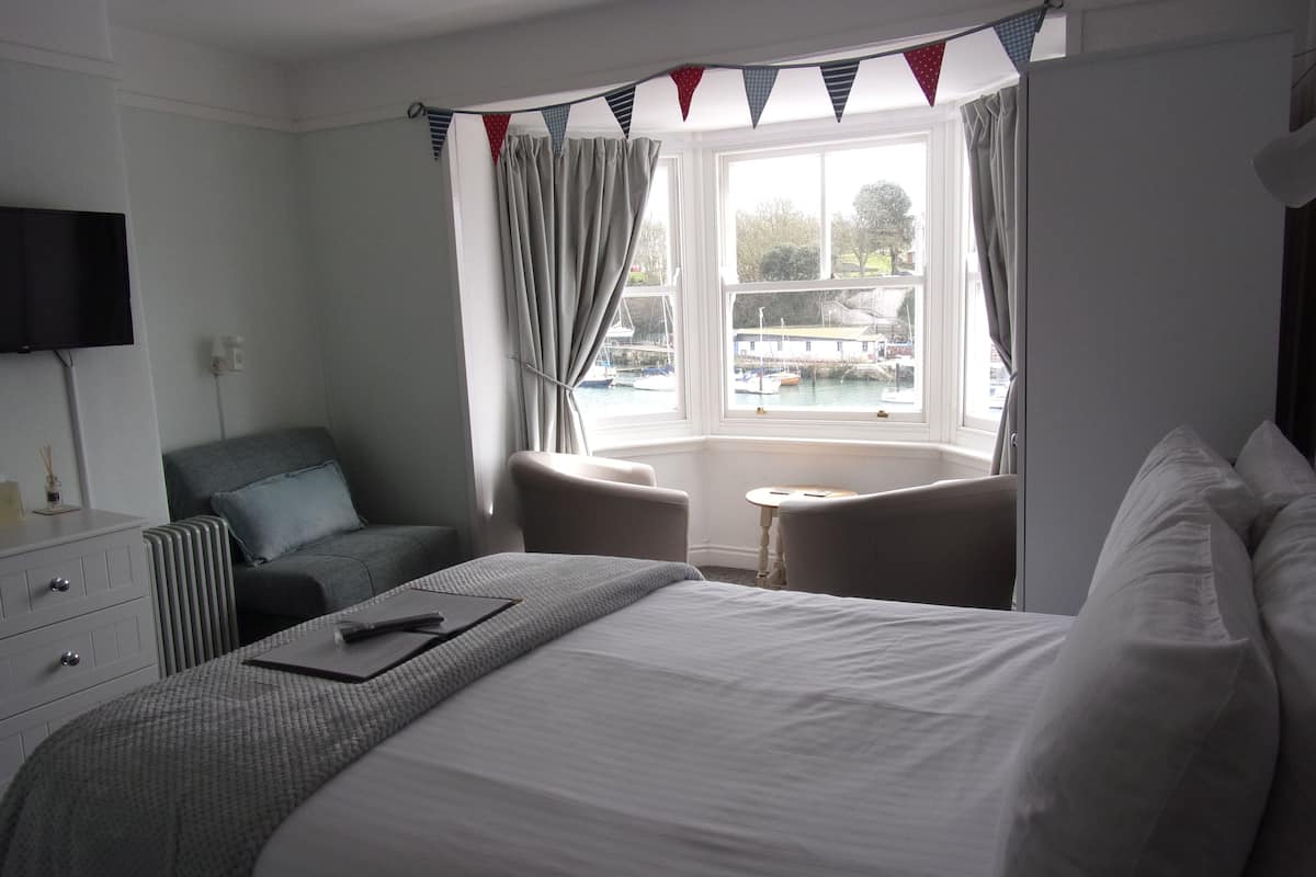 A hotel room with a large bed, a desk, a chair, and a view of boats through a window.