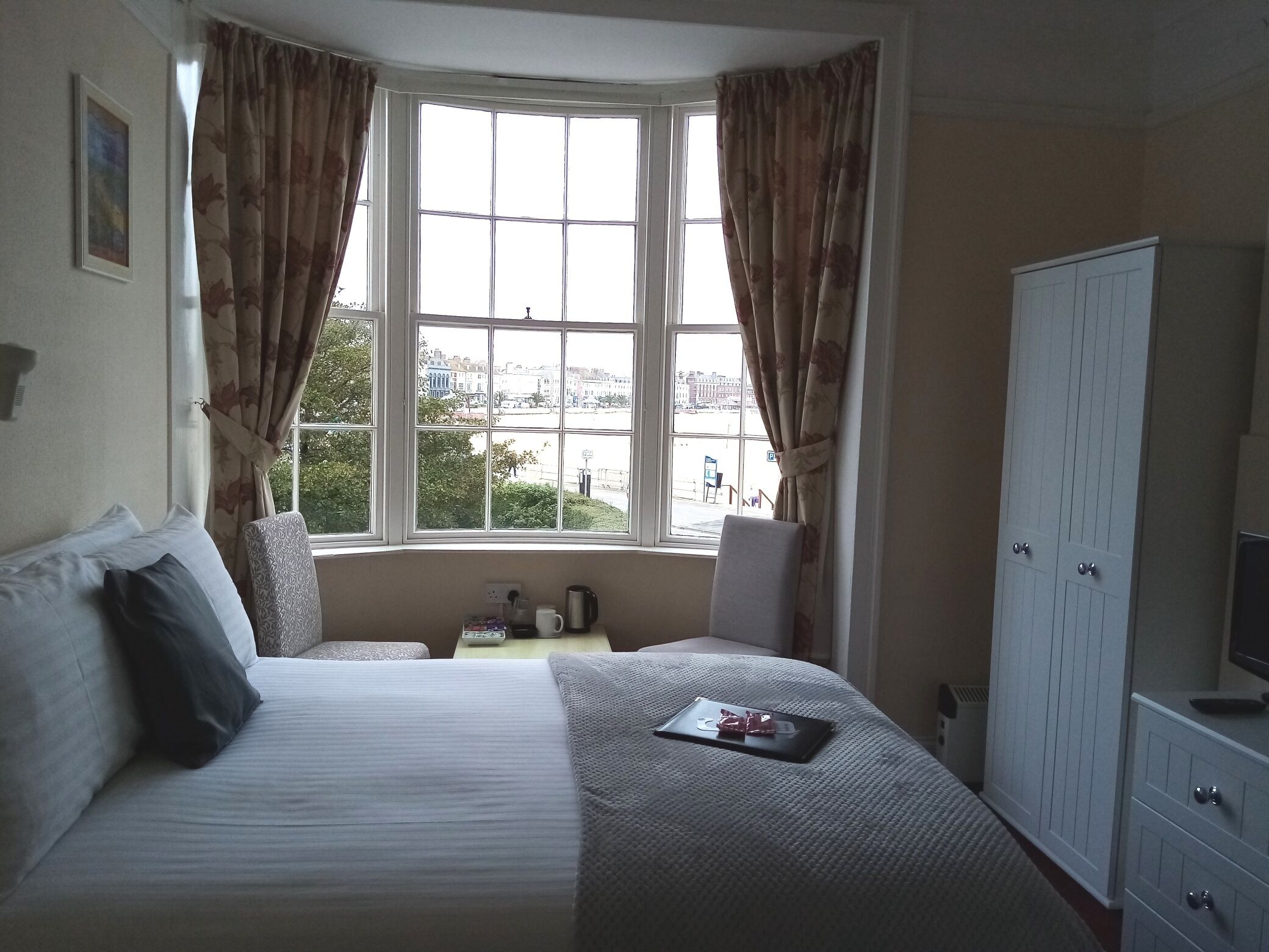 Une chambre d’hôtel avec une grande fenêtre, un lit, un bureau avec une chaise et une armoire.