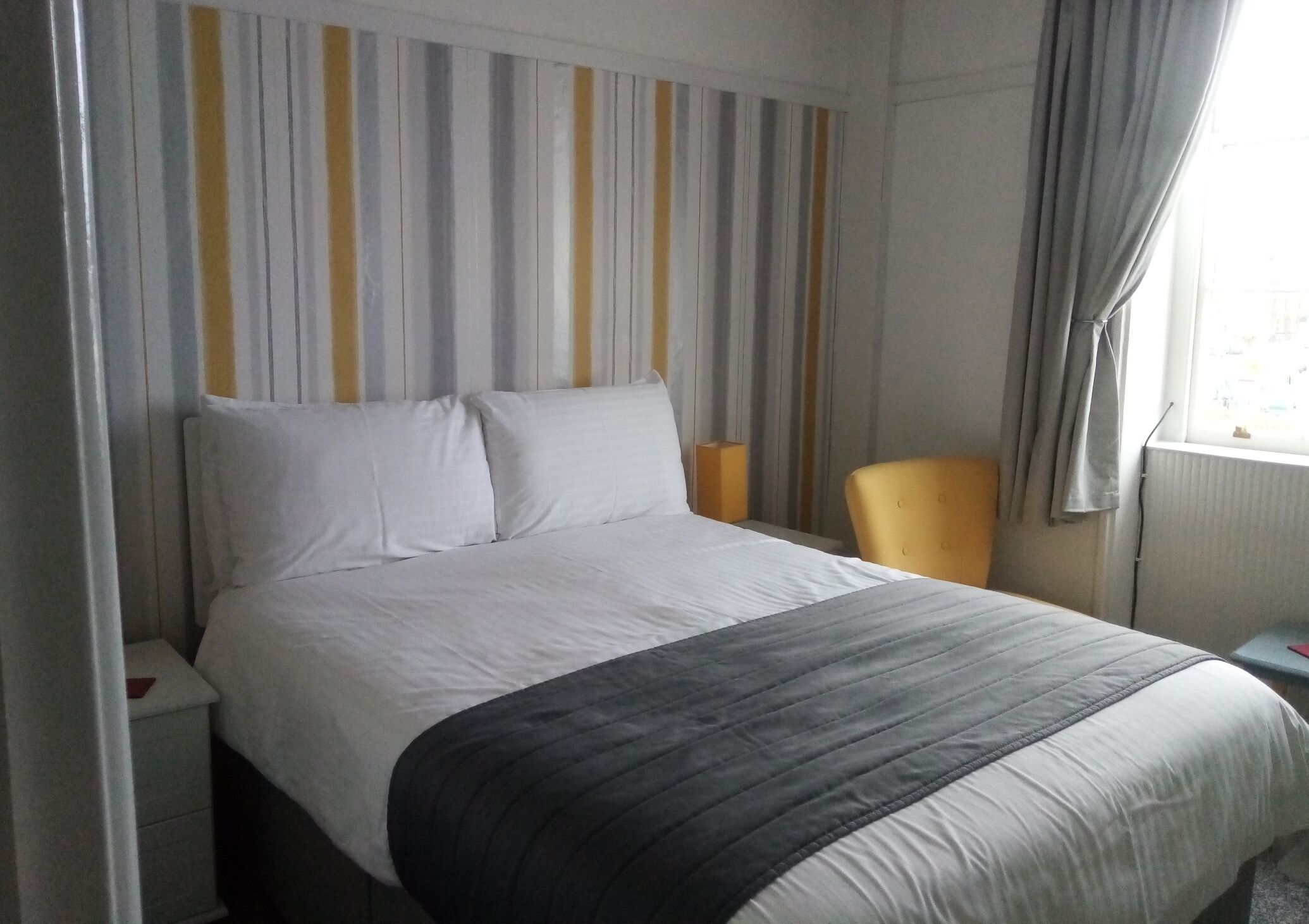 Ein Hotelzimmer mit einem Bett, einem Sessel, einem Nachttisch und einem Fenster mit Vorhängen.