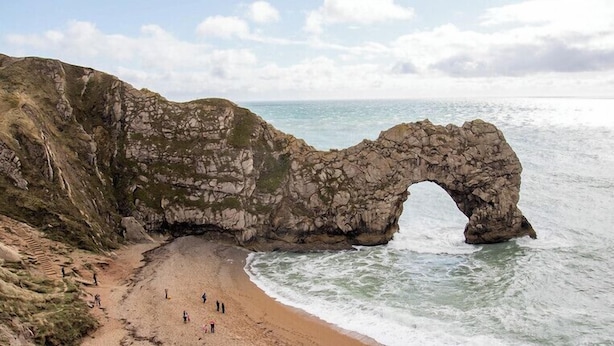 A coastal landscape with a prominent rock arch formation, a sandy beach, and a clear view of the sea. - The Cavendale (Weymouth)