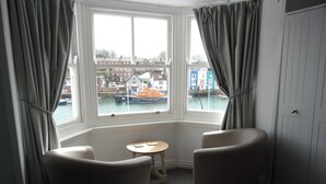 A room with two armchairs, a small table, and a view of a harbor with boats and colorful buildings. - The Cavendale (Weymouth)