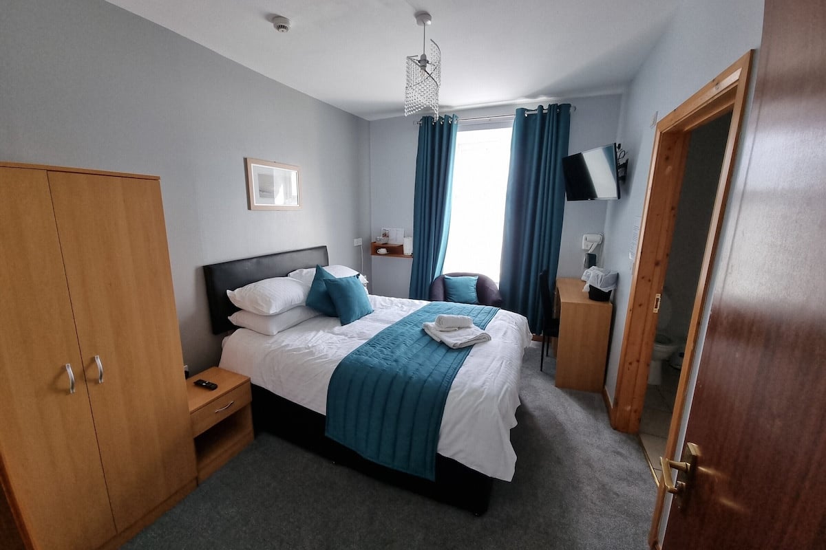 Ein Hotelzimmer mit Bett, Holzschrank, Nachttisch, Fernseher und einem Vorhangfenster.