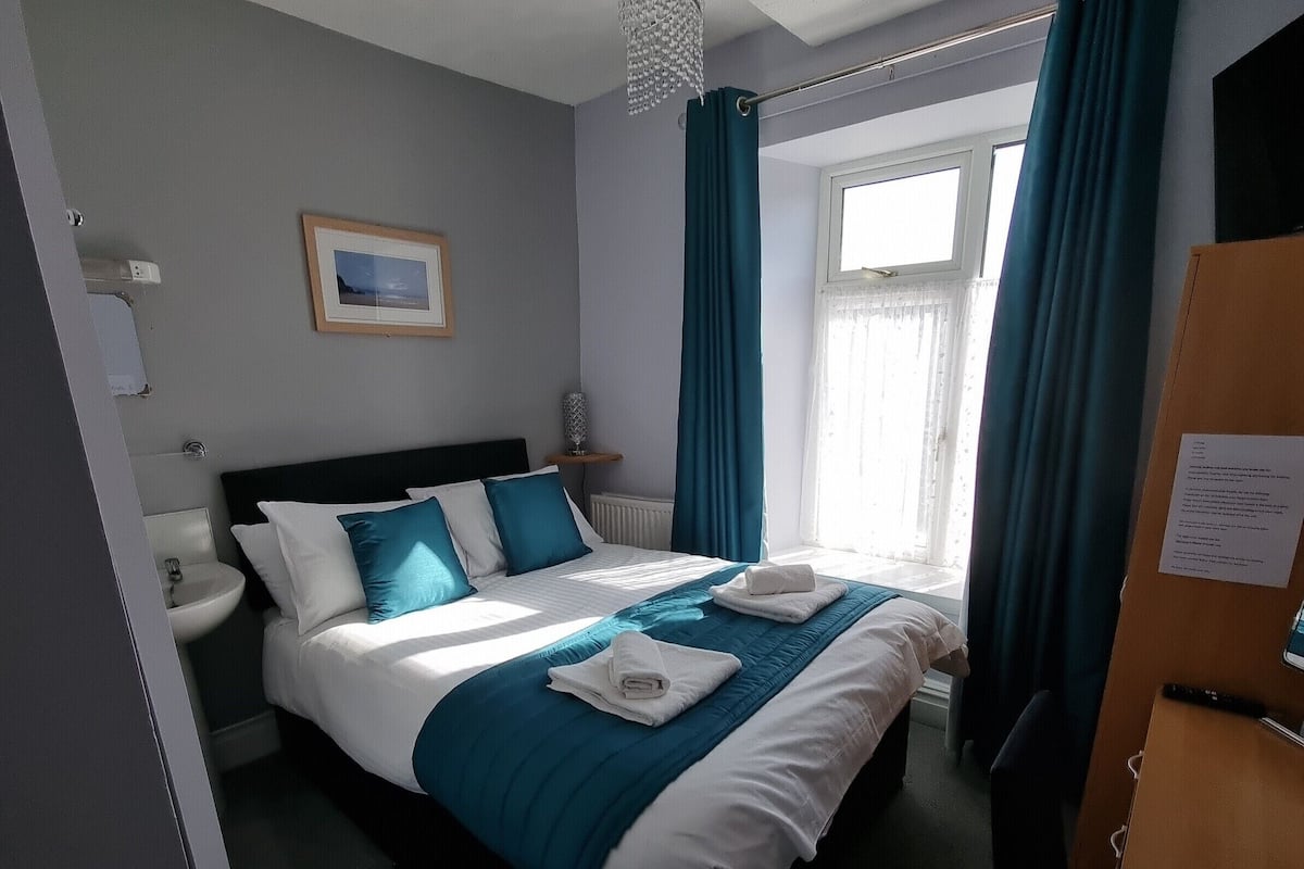 Ein Hotelzimmer mit Bett, Nachttisch, einem Fenster mit Vorhängen, einem Heizkörper und einem Bild an der Wand.
