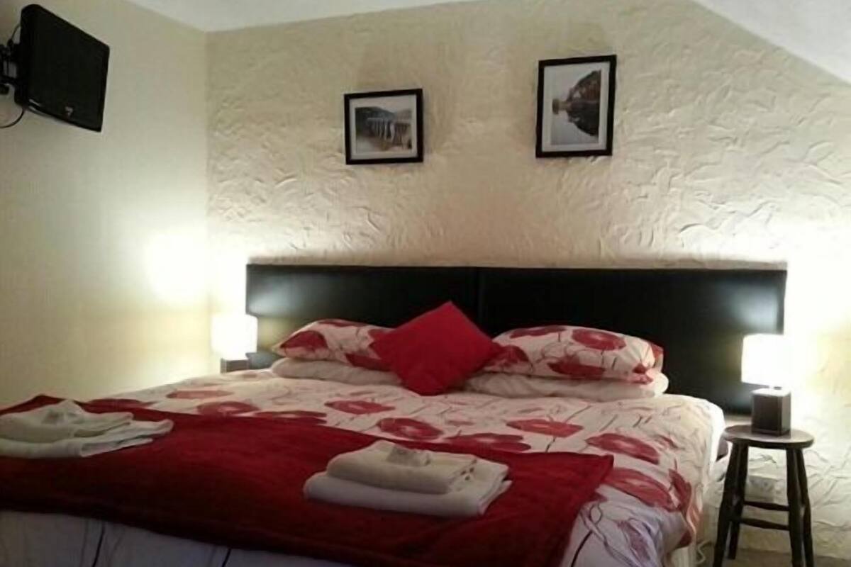 A bedroom with a bed, two wall-mounted pictures, a bedside table with a lamp, and a small stool.