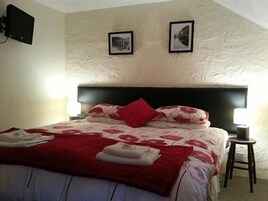 Quarto com cama, duas imagens penduradas na parede, mesa de cabeceira com abajur e um banquinho pequeno.