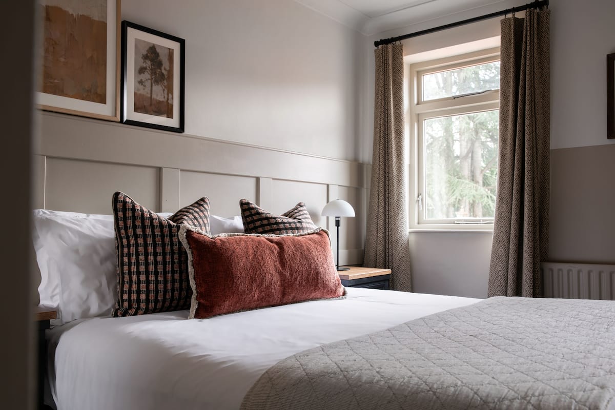 Una cama bien hecha con edredón blanco y varios cojines estampados, una mesita de noche con lámpara y una ventana con cortinas.