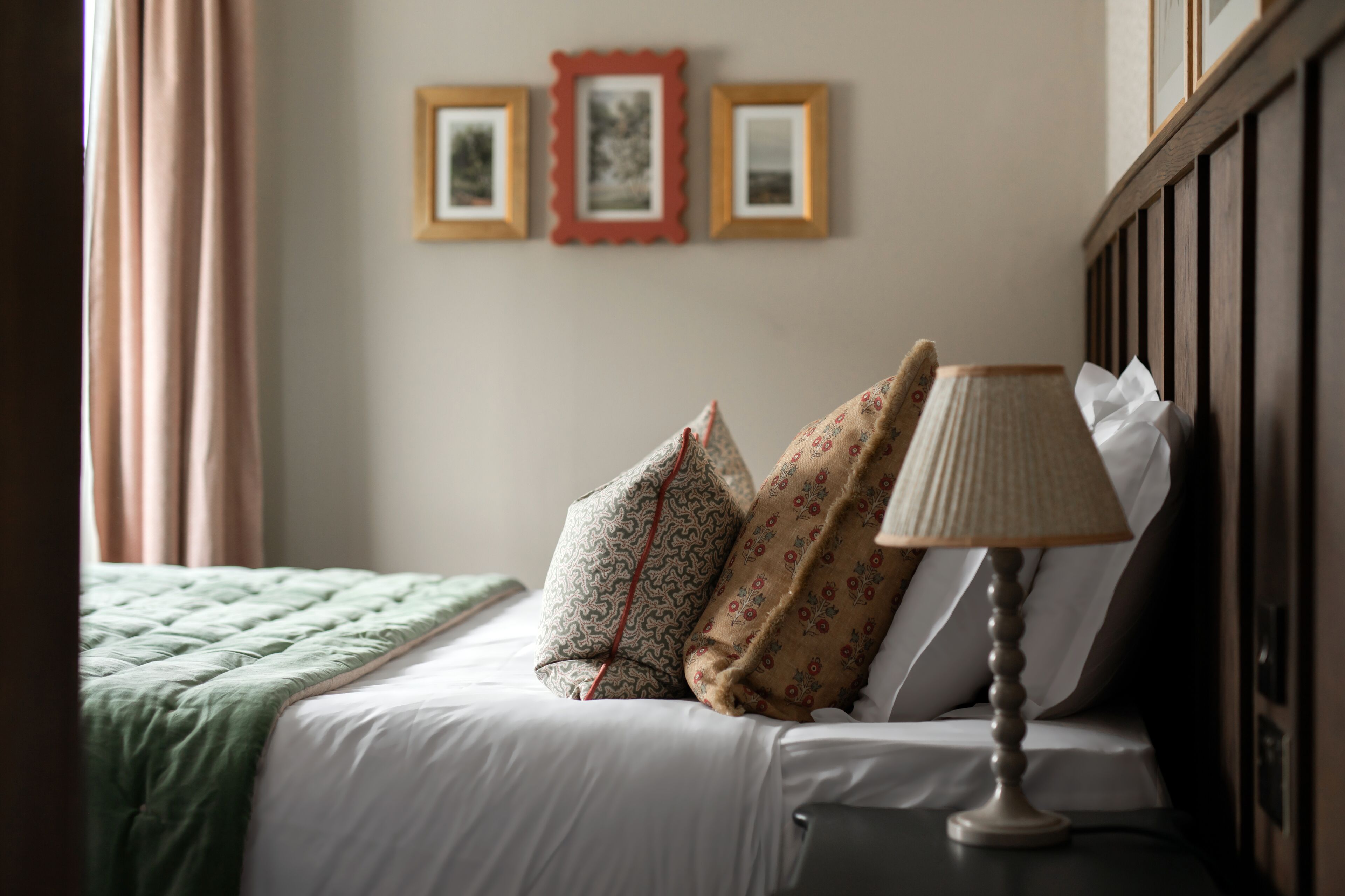 Una cama bien hecha con un edredón verde, almohadas estampadas y una lámpara de noche.