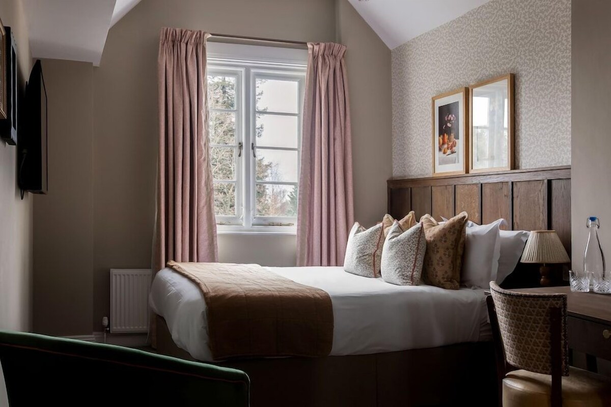 A hotel room with a bed, a window with curtains, a radiator, a wooden headboard, a chair, and a wall-mounted TV.