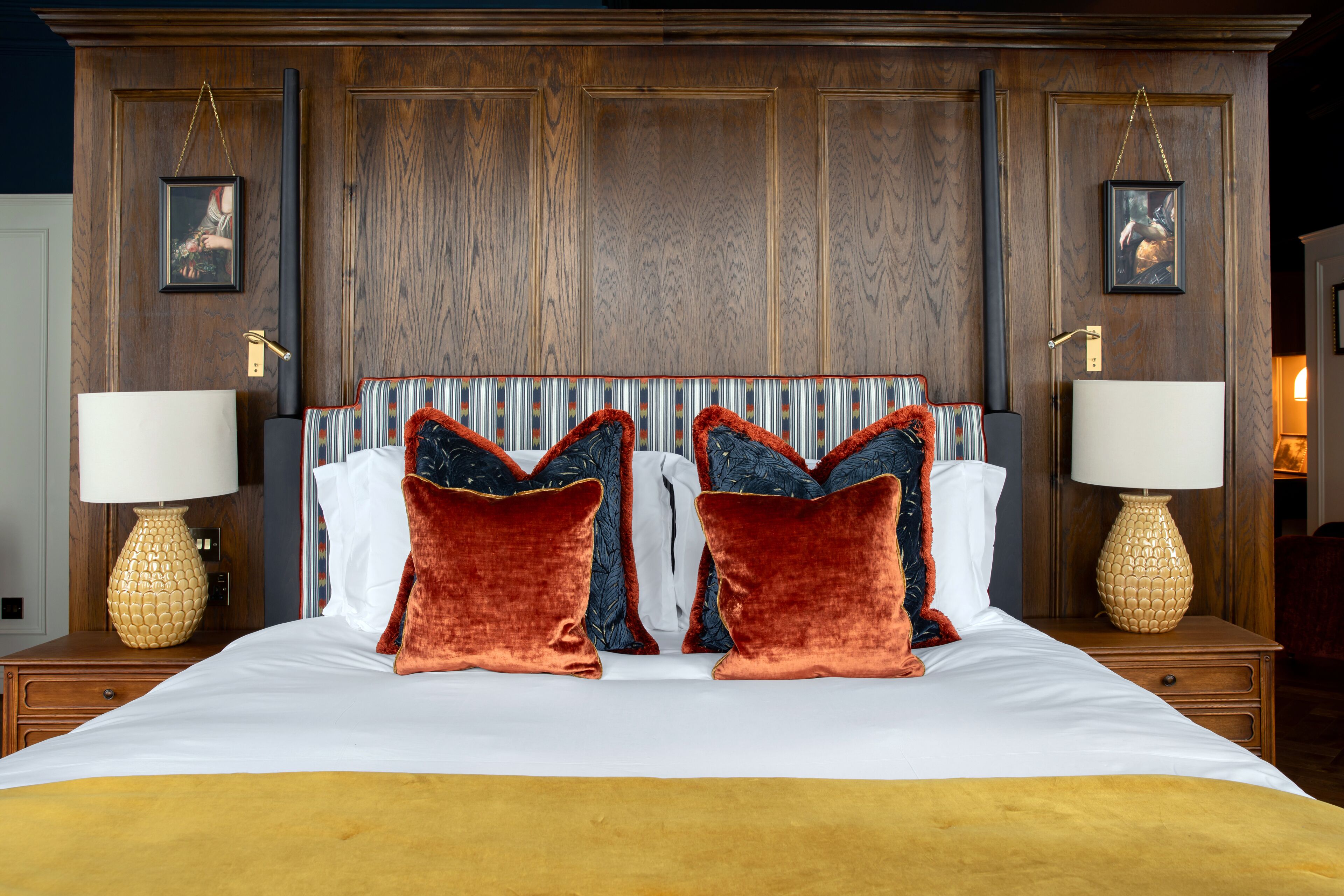 Una cama grande con cabecera de madera, dos mesitas de noche con lámparas y dos cuadros enmarcados en la pared.