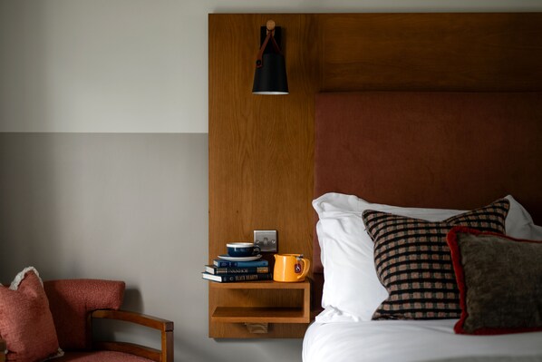 A hotel room with a wooden headboard, a bedside table with books and a mug, a chair, and a lamp.