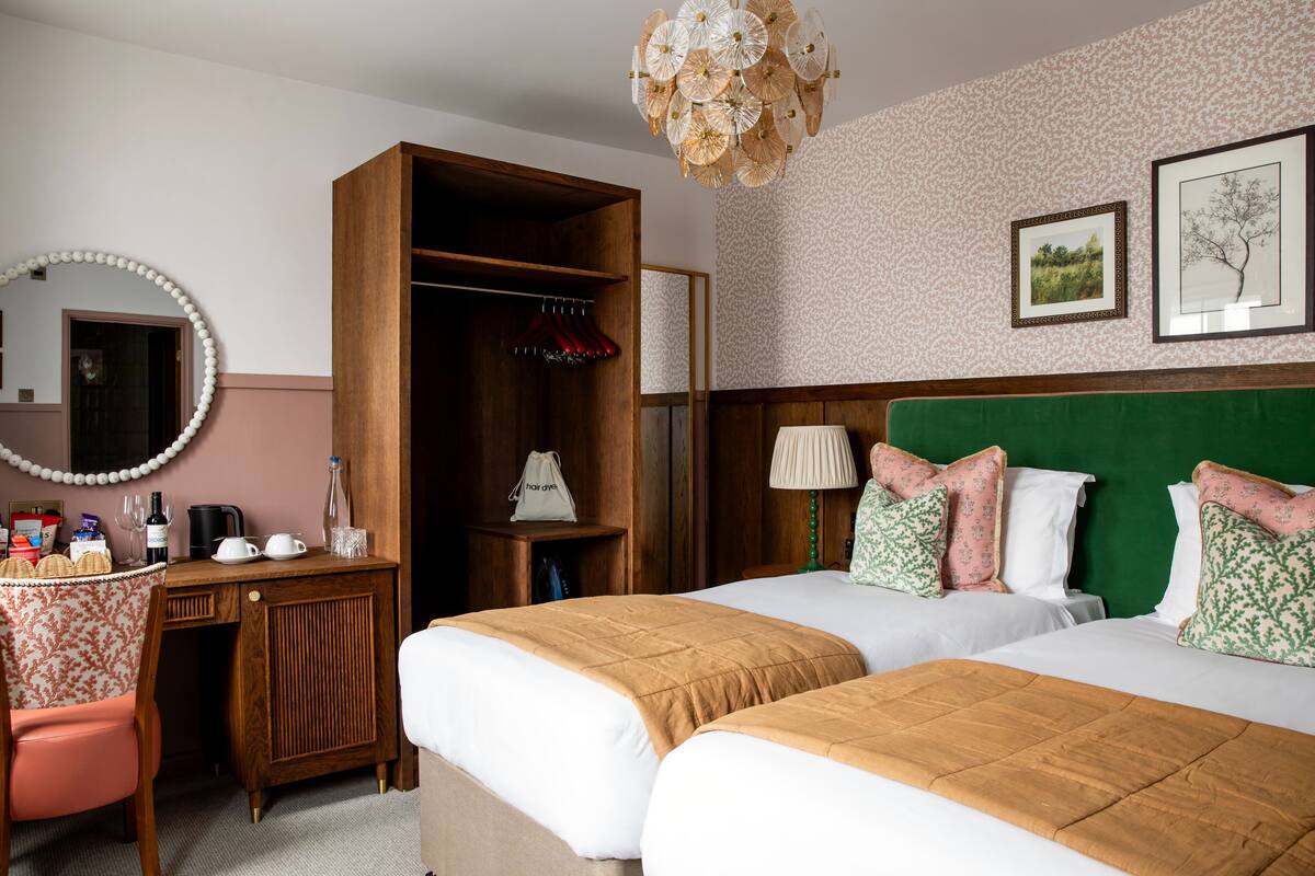 Una habitación de hotel con dos camas, un escritorio de madera, un espejo, un armario y una araña.
