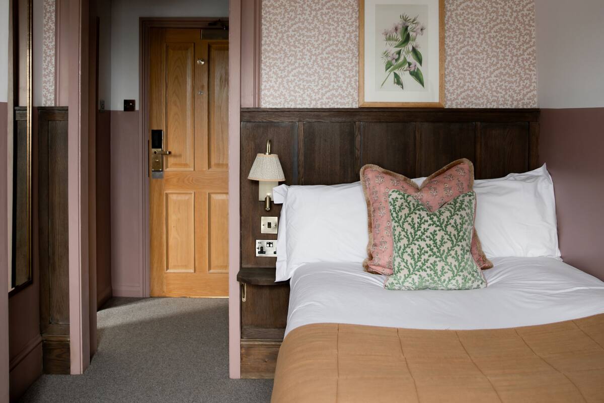 Una habitación de hotel con una cama grande, una puerta de madera, un cuadro enmarcado en la pared y una mesita de noche con una lámpara.