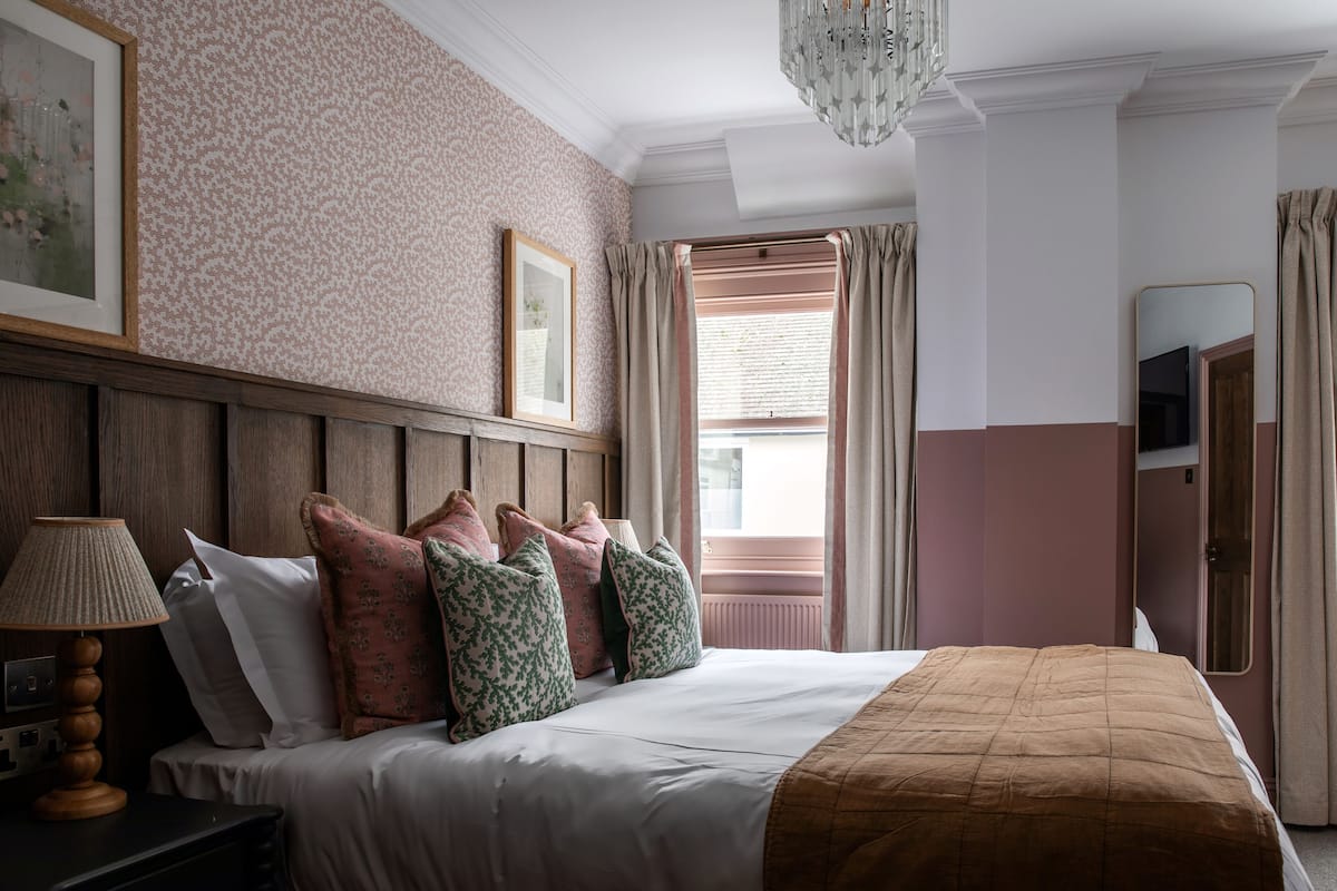 Una habitación de hotel con una cama grande, cabecera de madera, un cuadro enmarcado, una lámpara y una ventana con cortinas.