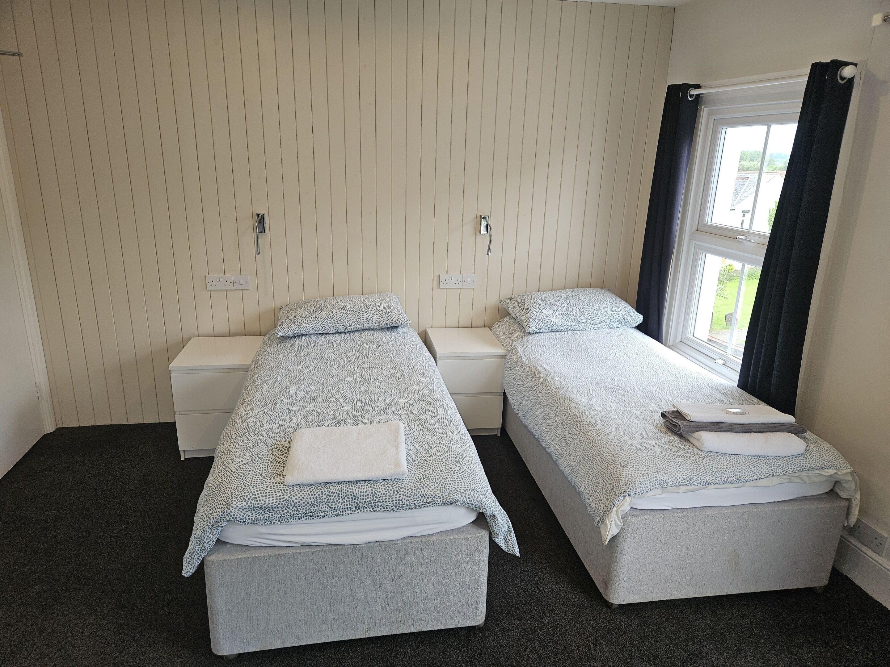 A room with two single beds, each with a matching nightstand and a bedside table. The walls are paneled, and there is a window with curtains.