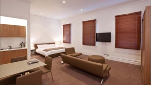 Quarto de hotel com cama, escrivaninha, poltrona, sofá, televisão e persianas de madeira.