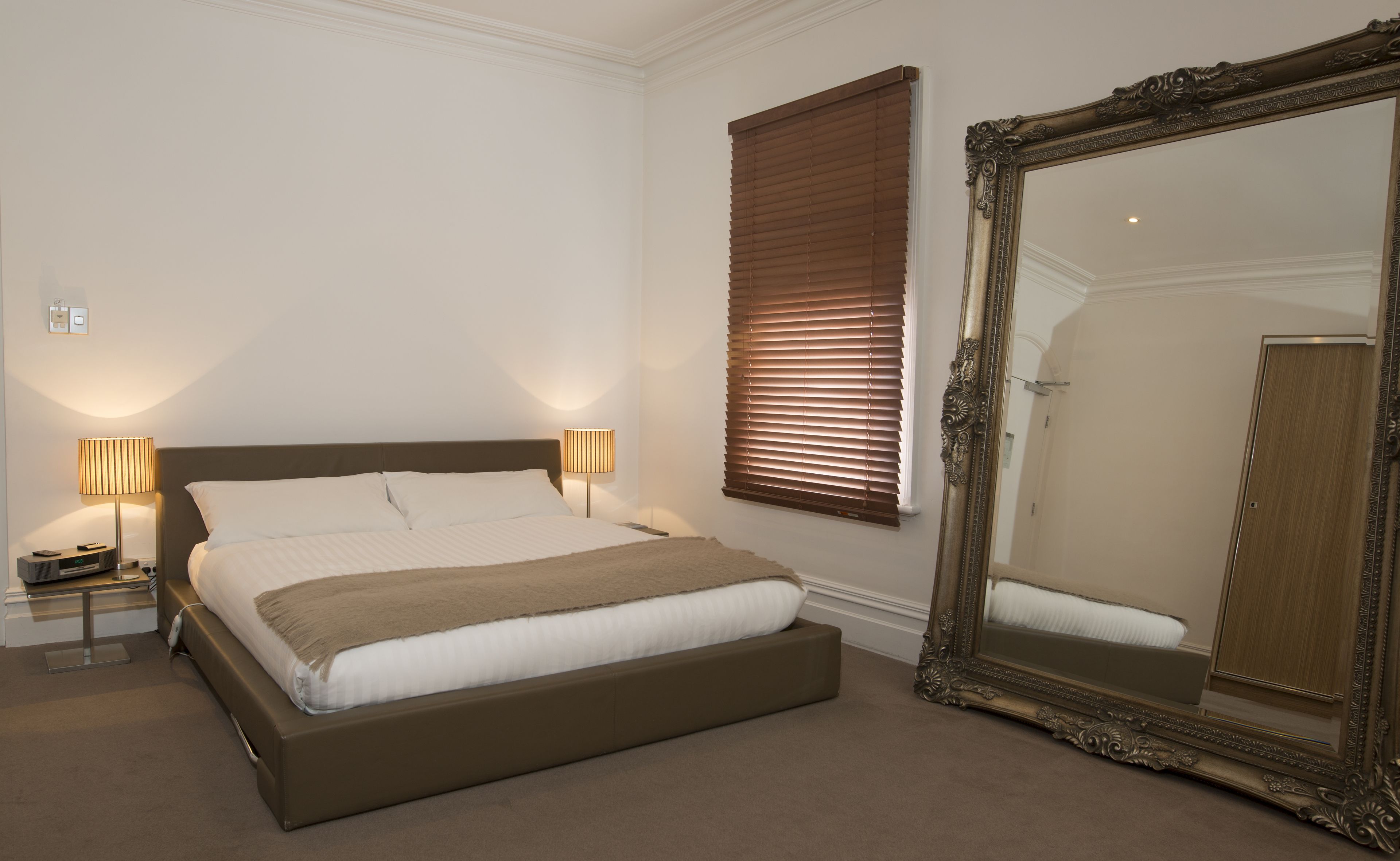 A hotel room with a large bed, two bedside lamps, a window with blinds, and a large ornate mirror.