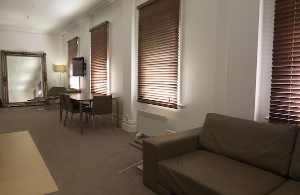 A modern living room with a sofa, dining table, chairs, and large windows with blinds.