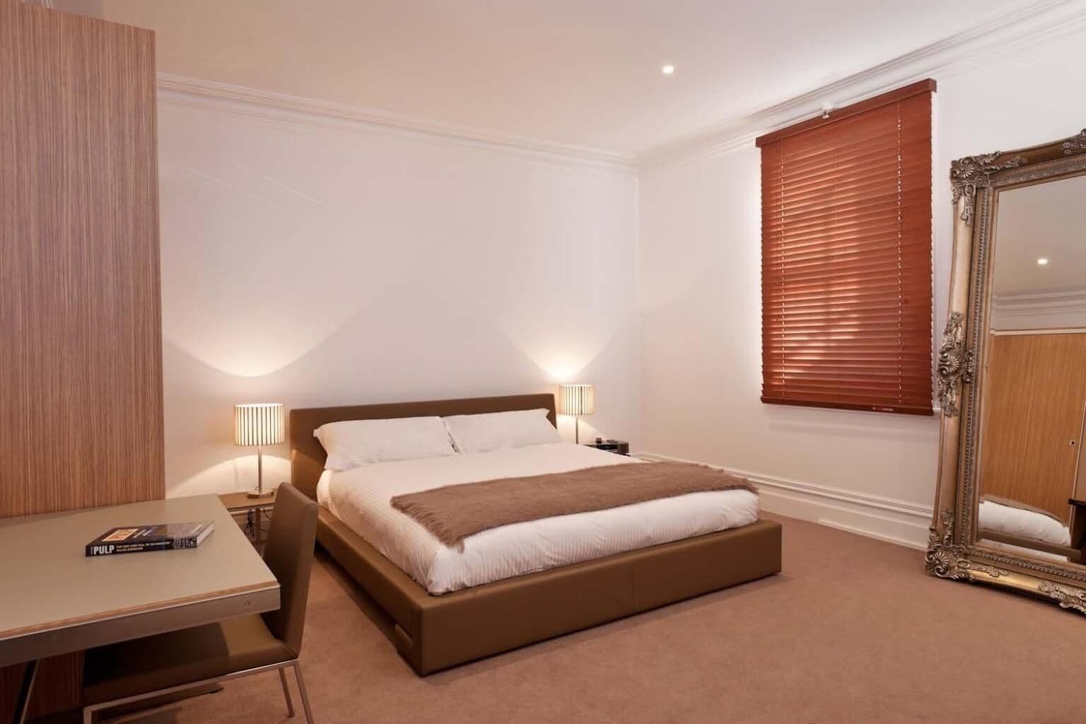 Quarto de hotel com cama, luminárias de cabeceira, escrivaninha e um espelho grande.