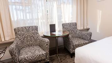 Ein Hotelzimmer mit zwei gemusterten Sesseln, einem kleinen Glastisch, einem Bett und Blick ins Freie durch transparente Vorhänge.