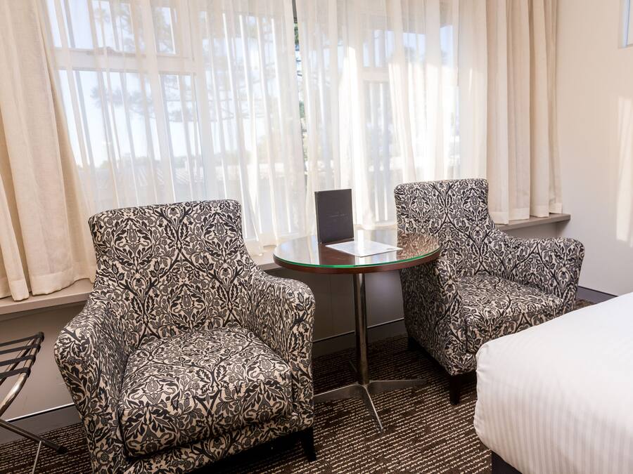 Ein Hotelzimmer mit zwei gemusterten Sesseln, einem kleinen Glastisch, einem Bett und Blick ins Freie durch transparente Vorhänge.