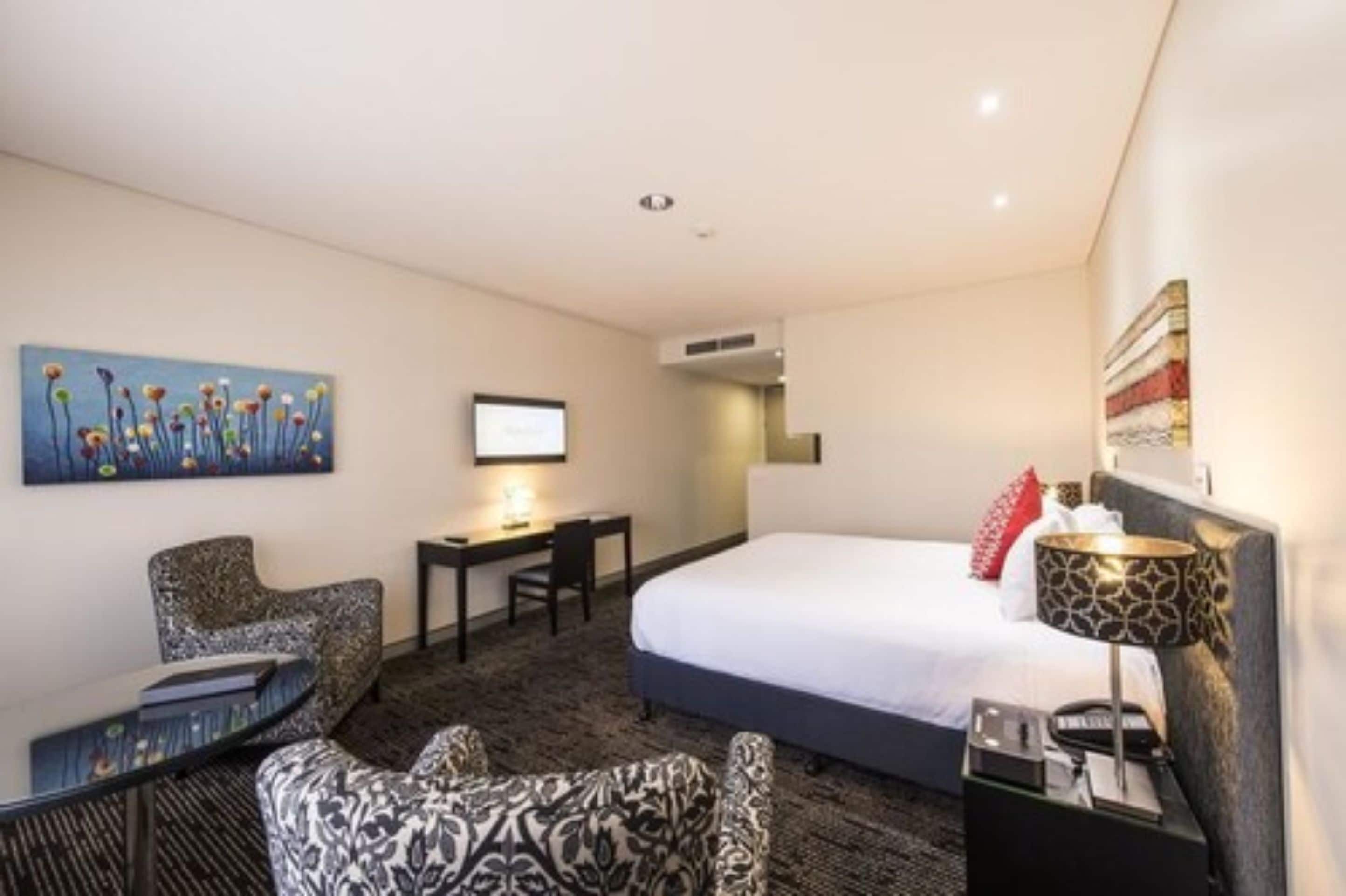 A hotel room with a bed, a desk, a chair, a small table, and a painting on the wall.
