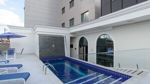 Une piscine aux carreaux bleus, un espace couvert avec des chaises et un parasol, et un bâtiment à la façade carrelée.