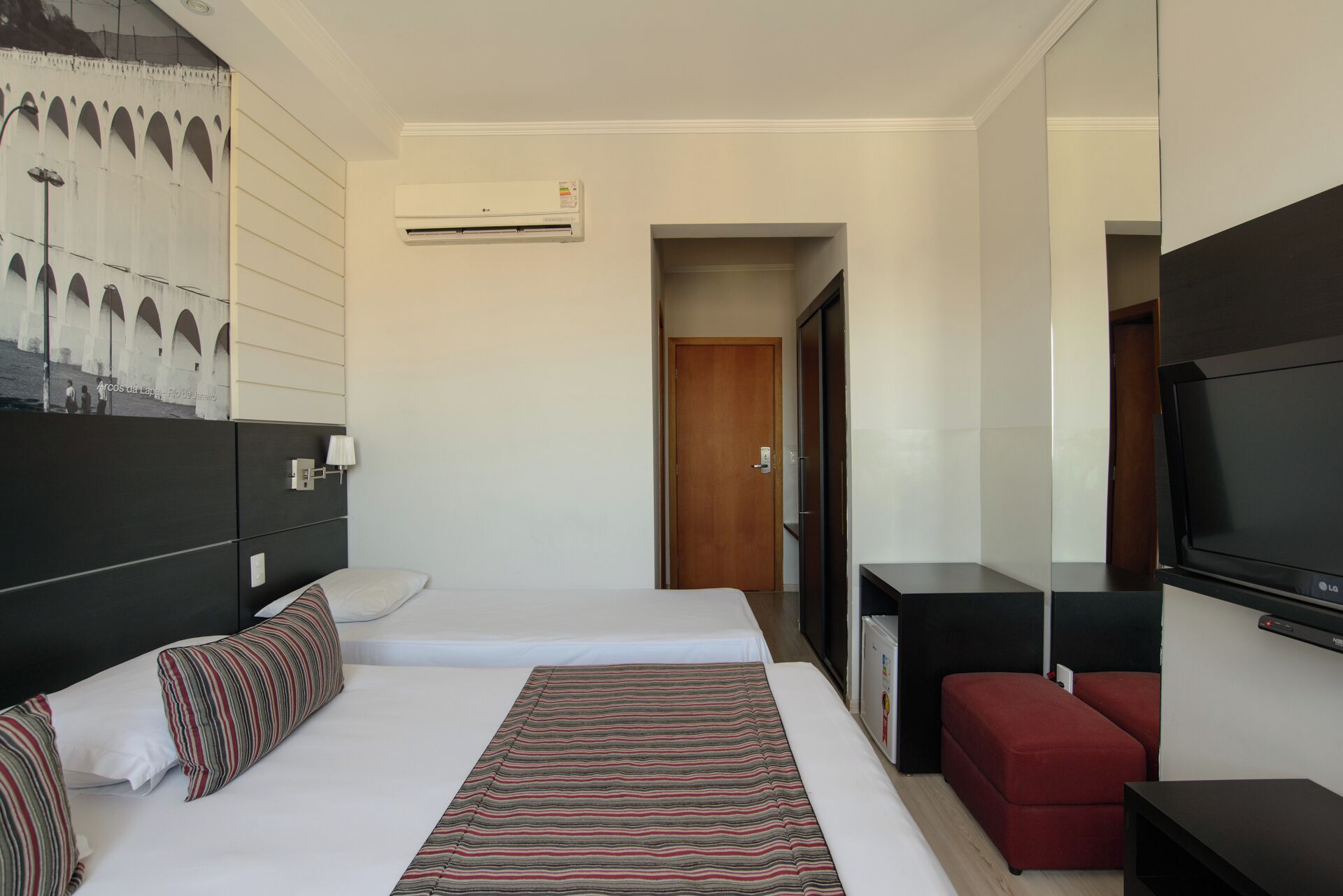 Quarto de hotel com duas camas, uma televisão, uma escrivaninha e um sofá vermelho.
