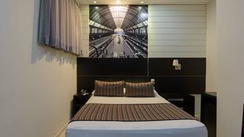 Quarto de hotel com uma cama grande, mesa de cabeceira com telefone, luminária na parede e vista para uma estação de trem.