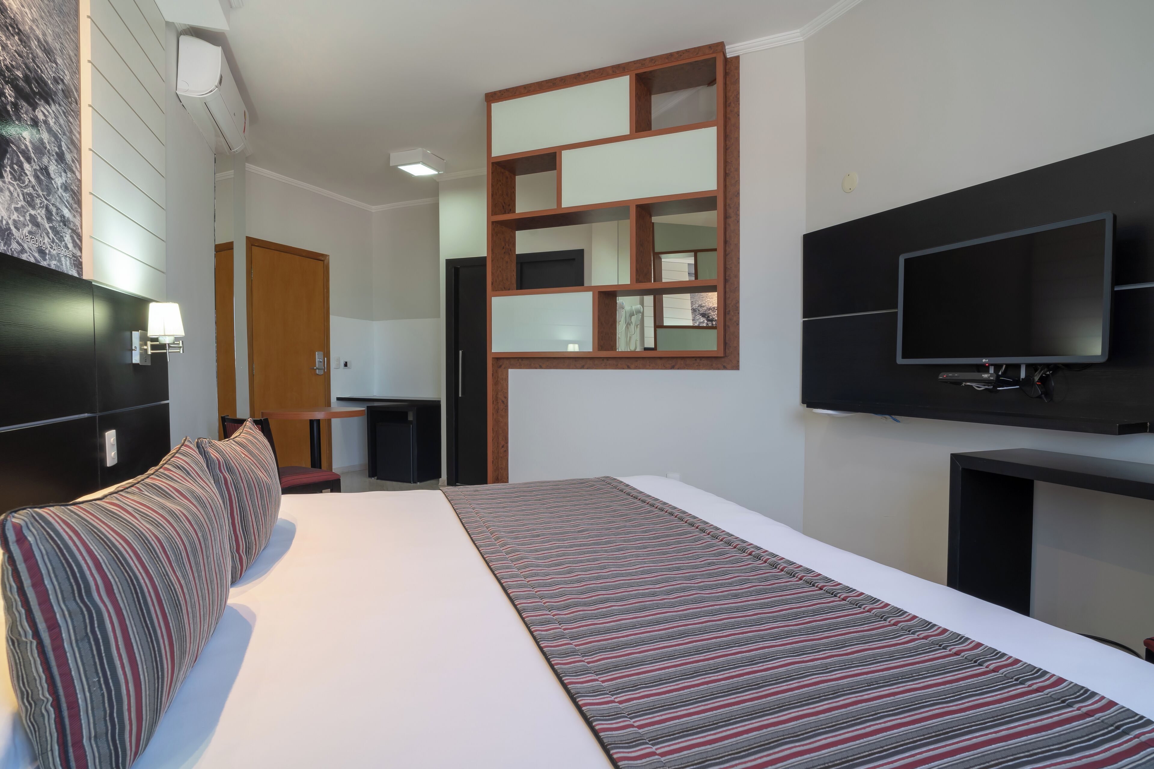 A modern hotel room with a large bed, a flat-screen TV, a wooden-framed mirror, and a small desk.