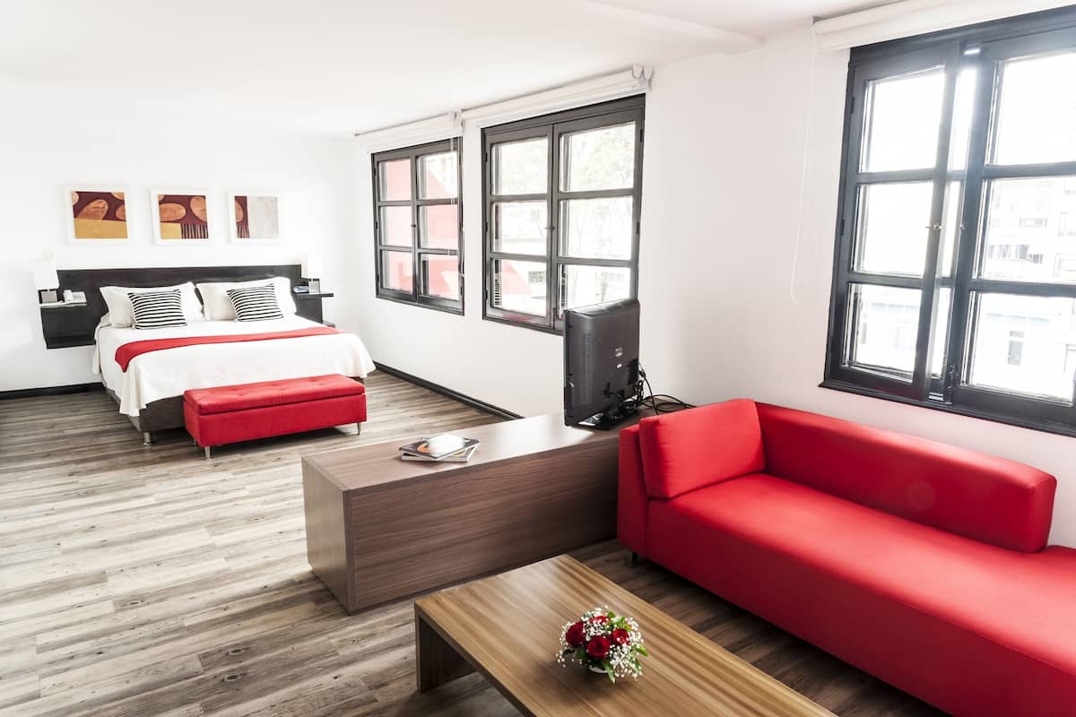 Camera d'albergo moderna con un letto, un divano rosso, un tavolino in legno e ampie finestre.