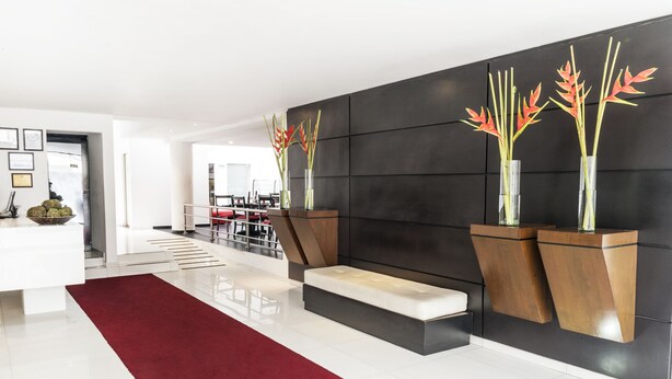 Un vestíbulo moderno con alfombra roja, un mostrador de recepción blanco y dos jarrones con plantas sobre una pared oscura.